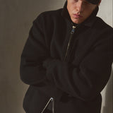 A model wearing a black wool zip-up jacket, black baseball cap, and black pants, standing with arms crossed against a textured gray wall.