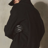 A model wearing a black wool coat, black leather gloves, and a black cap, shown from a side-back angle against a textured concrete wall.
