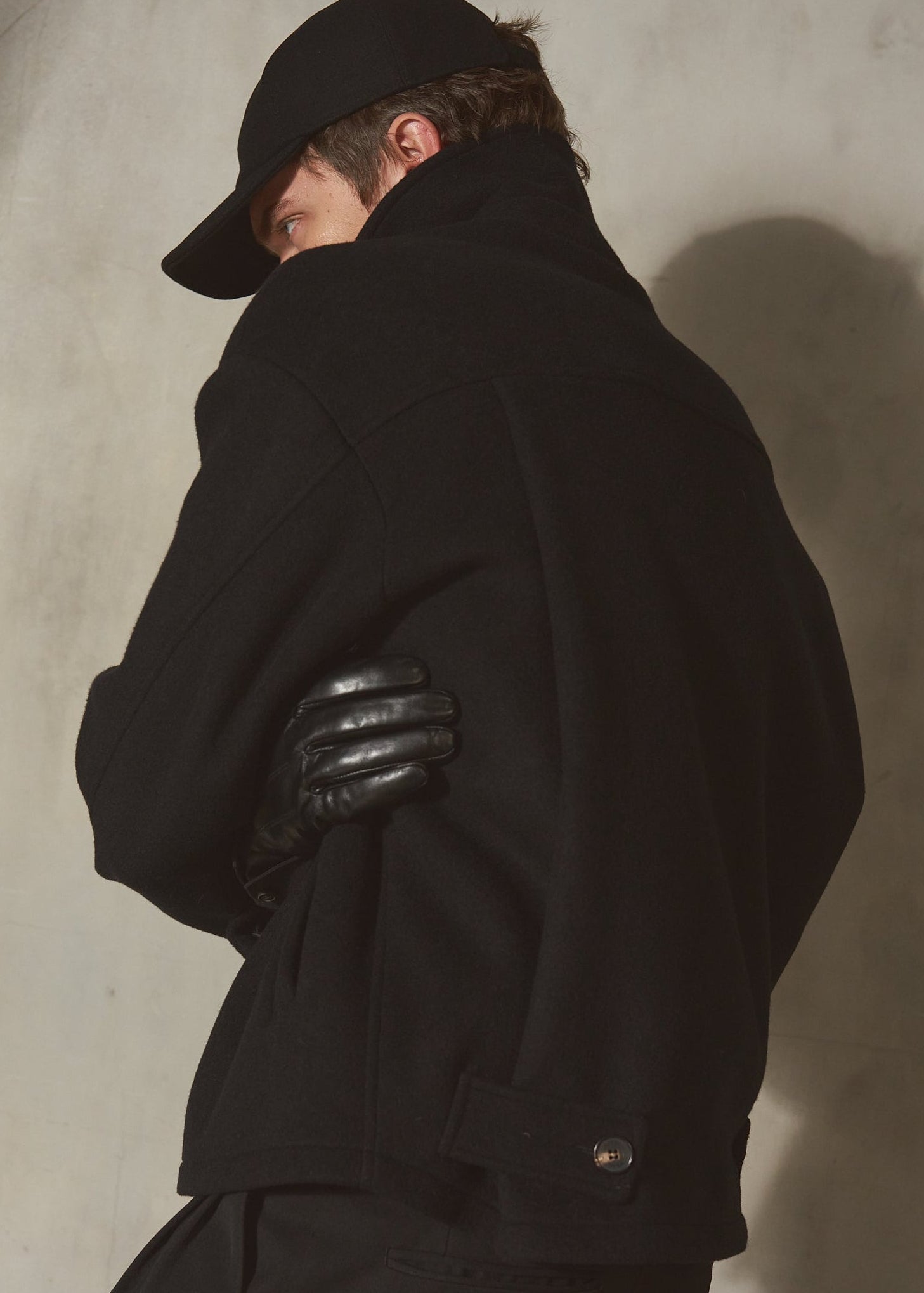 A model wearing a black wool coat, black leather gloves, and a black cap, shown from a side-back angle against a textured concrete wall.