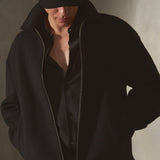 A man wearing a black zip-up wool jacket, black wool baseball cap, and black shirt, standing against a textured gray wall.