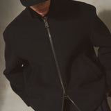 A person wearing a black wool zip-front jacket with a high collar, matching black pants, and a black cap, photographed against a textured light gray wall.