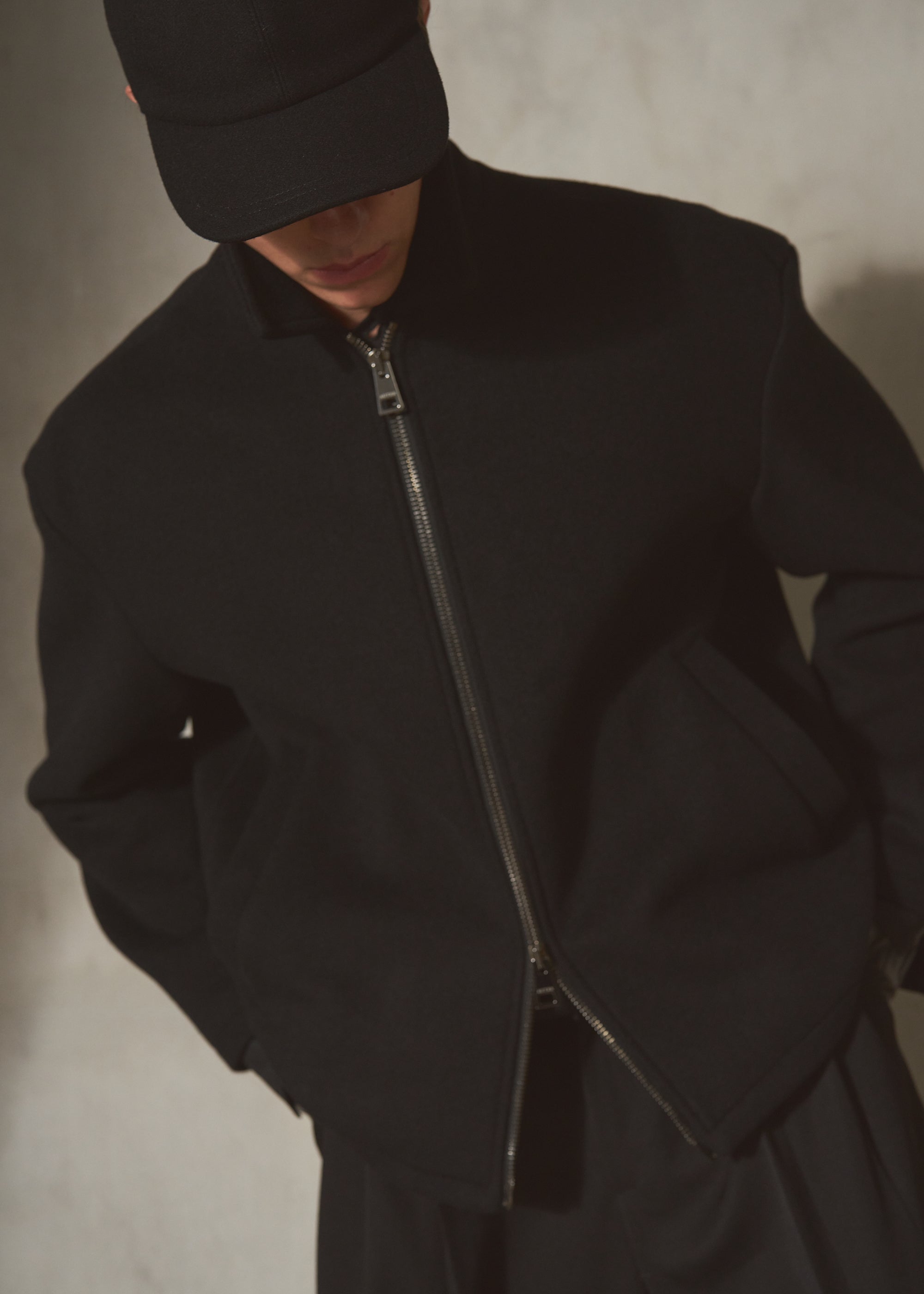 A person wearing a black wool zip-front jacket with a high collar, matching black pants, and a black cap, photographed against a textured light gray wall.