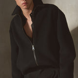 A man wearing a wool black zip-up jacket, black cap, black pants, and black gloves, standing against a textured gray wall with dramatic side lighting.