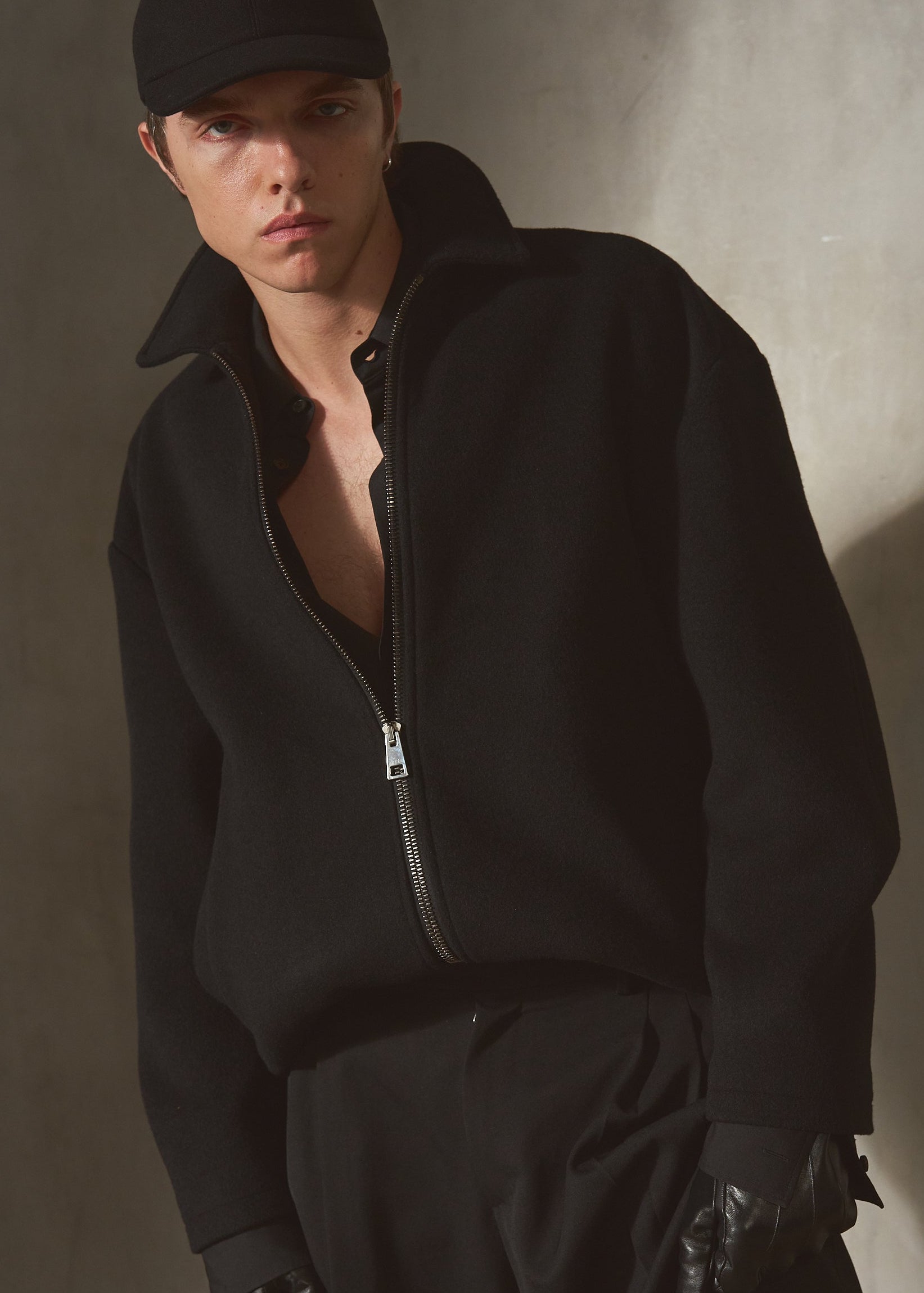 A man wearing a wool black zip-up jacket, black cap, black pants, and black gloves, standing against a textured gray wall with dramatic side lighting.