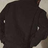 A man viewed from behind wearing a black wool jacket and matching black trousers, standing against a textured light gray wall.