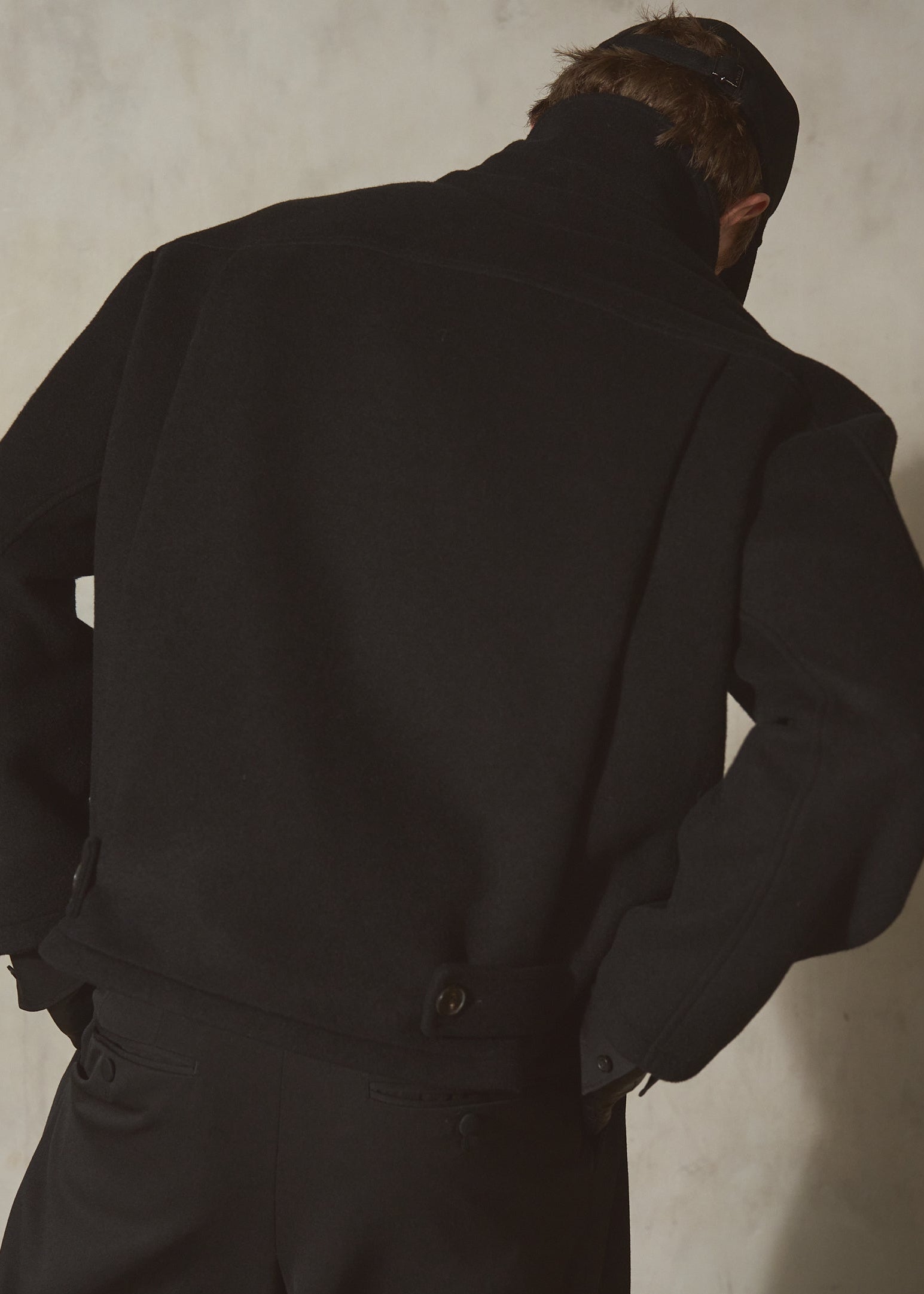 A man viewed from behind wearing a black wool jacket and matching black trousers, standing against a textured light gray wall.