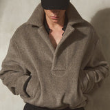 A male model wearing a grey herringbone knit pullover, black wide-leg trousers, and a black baseball cap stands against a textured concrete wall.