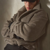 A model wearing an oversized taupe-brown textured knit sweater, a black baseball cap, and black trousers, leaning against a textured concrete wall with dramatic side lighting.