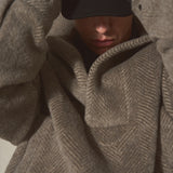 A close-up of a person wearing an oversized taupe-brown herringbone knit pullover, with a black cap pulled down over the eyes.