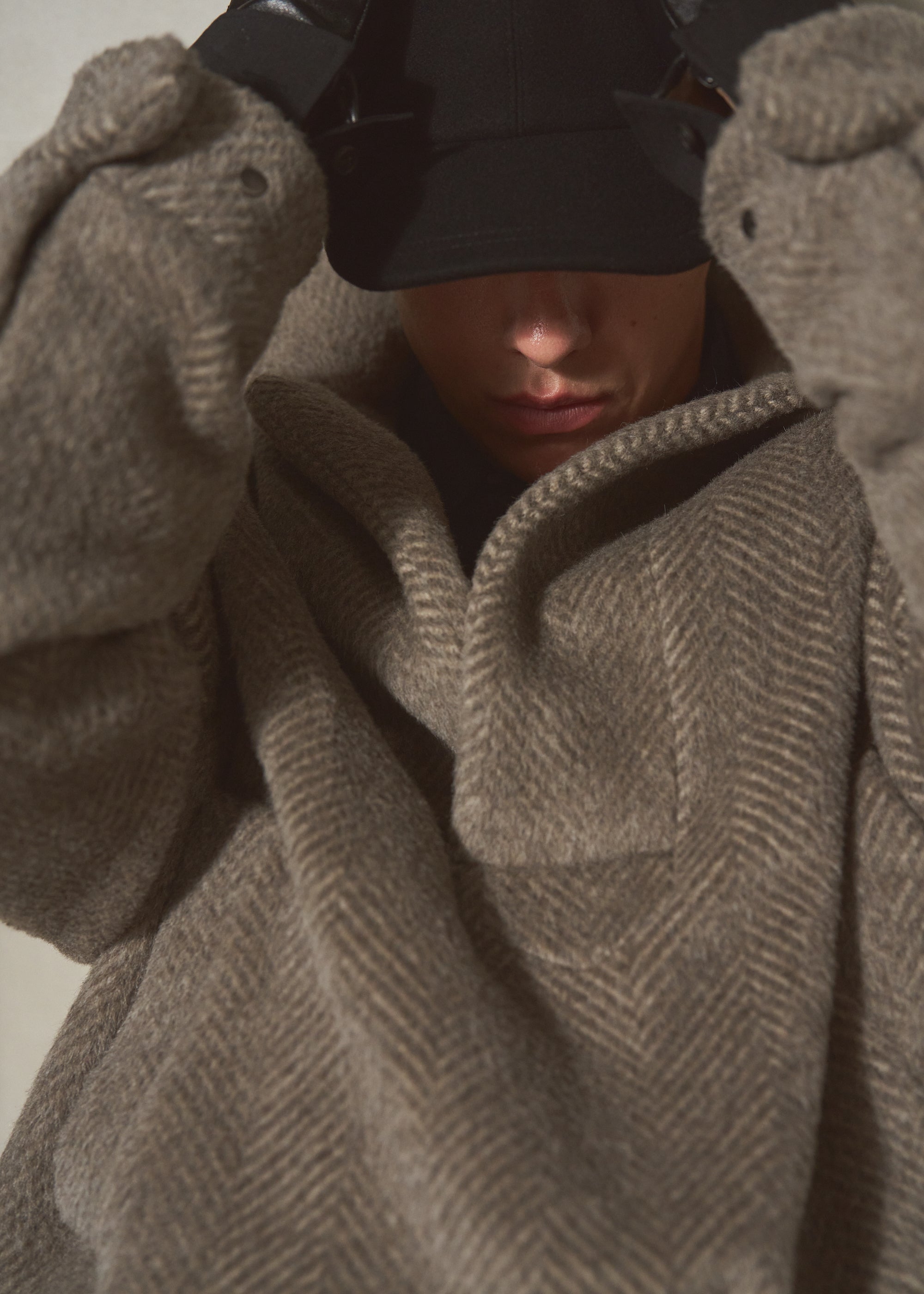 A close-up of a person wearing an oversized taupe-brown herringbone knit pullover, with a black cap pulled down over the eyes.