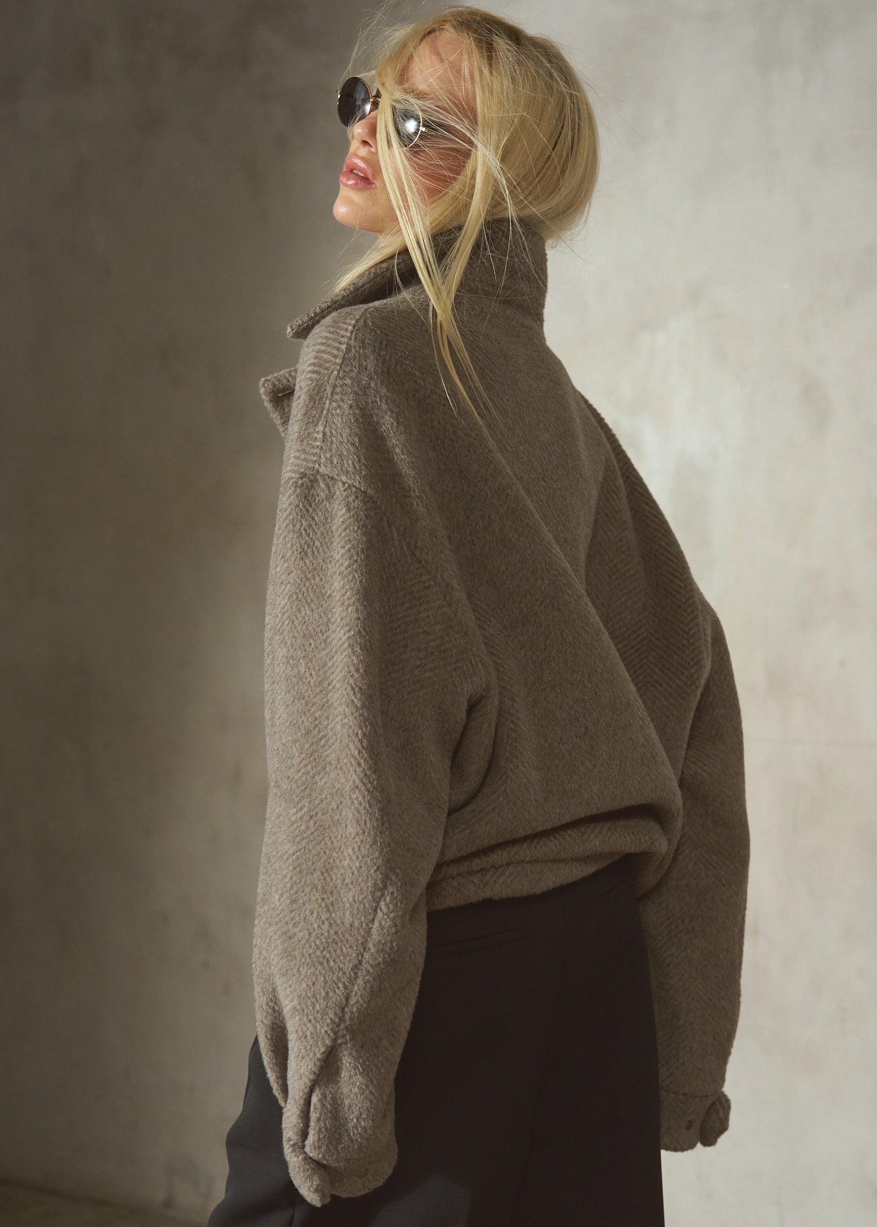 A woman wearing a loose, oversized taupe-brown herringbone pullover jacket and black wide-leg pants.