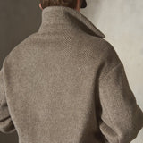 A model viewed from behind wearing a textured herringbone wool pullover, a black baseball cap, and dark trousers, set against a neutral gray studio background.
