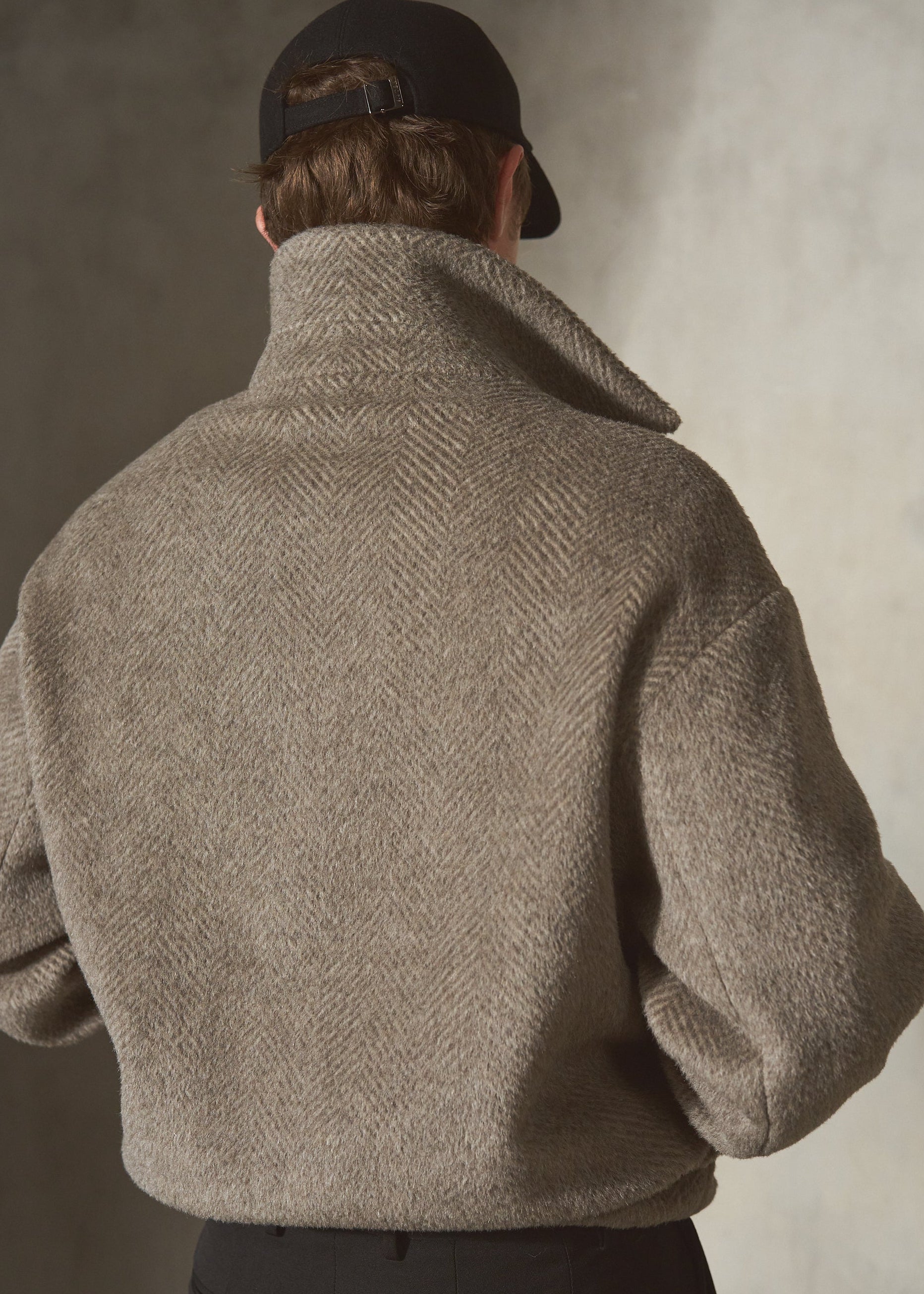 A model viewed from behind wearing a textured herringbone wool pullover, a black baseball cap, and dark trousers, set against a neutral gray studio background.