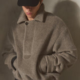 A model wearing a textured taupe-gray herringbone knit sweater with a high collar, a black baseball cap, and dark trousers, standing against a textured concrete wall with dramatic side lighting.