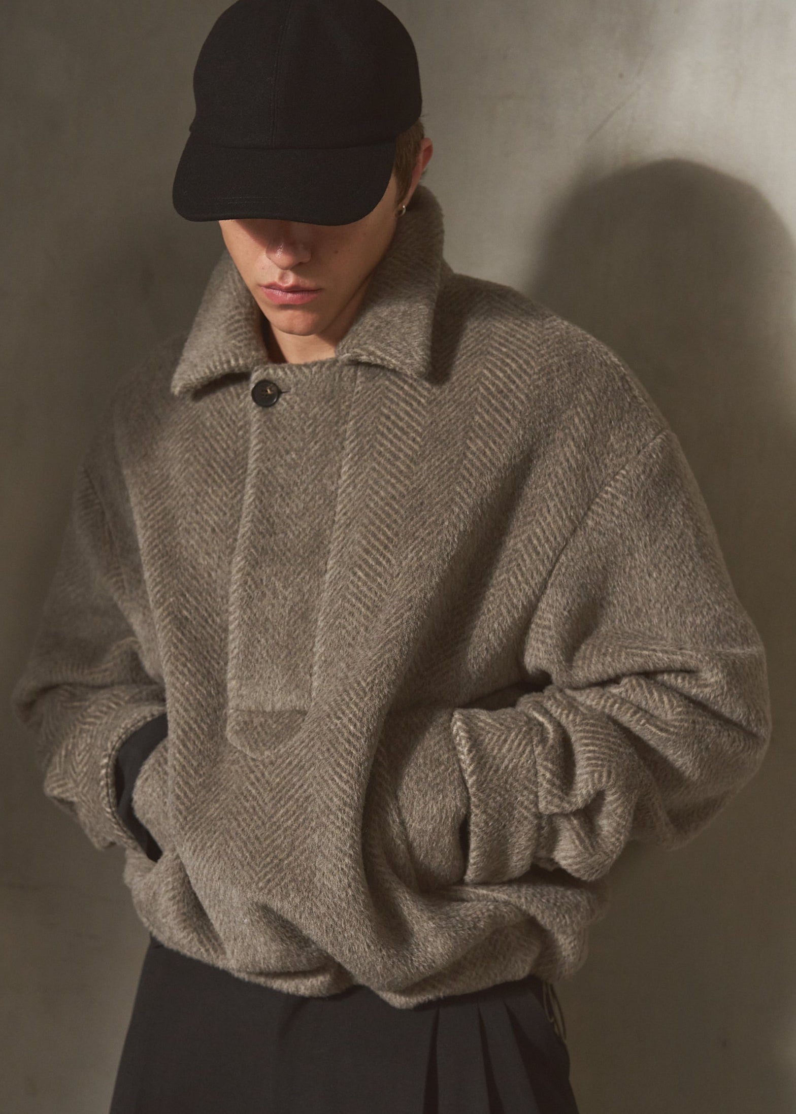A model wearing a textured taupe-gray herringbone knit sweater with a high collar, a black baseball cap, and dark trousers, standing against a textured concrete wall with dramatic side lighting.