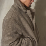 A model wearing a black baseball cap and a textured taupe-grey herringbone pullover, posed in profile against a neutral concrete wall.