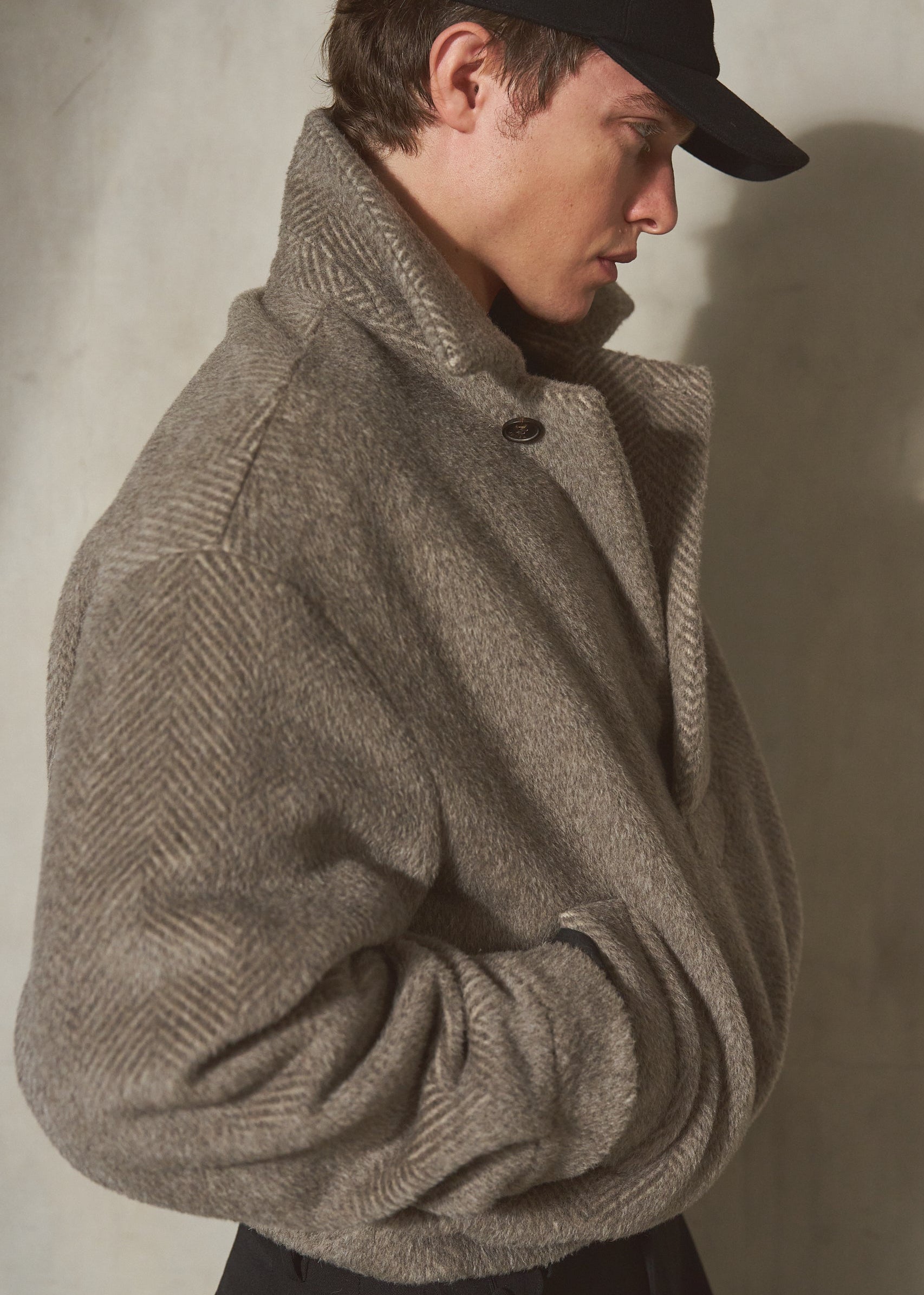 A model wearing a black baseball cap and a textured taupe-grey herringbone pullover, posed in profile against a neutral concrete wall.