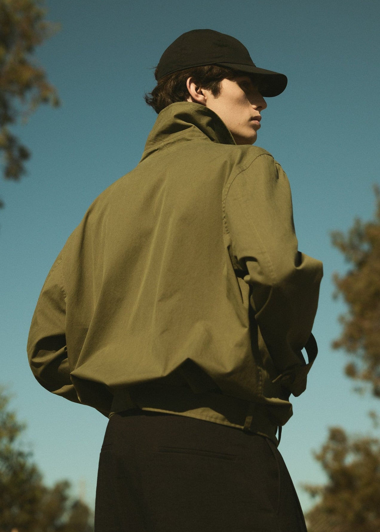 Person wearing a green jacket and black cap outdoors with trees in the background