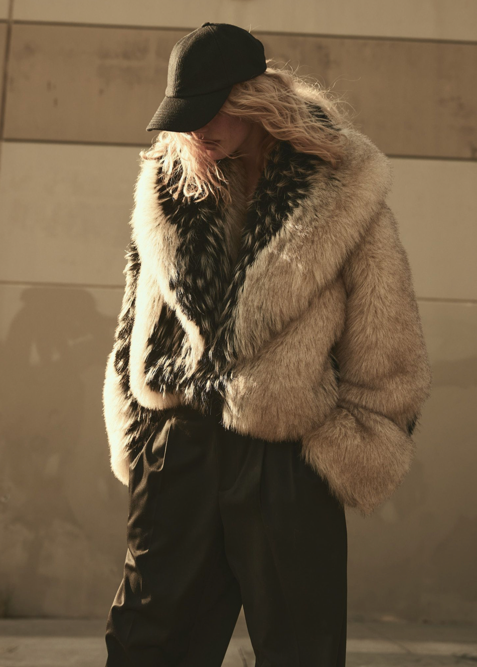 A woman wearing a faux fur coat and cap against a plain wall
